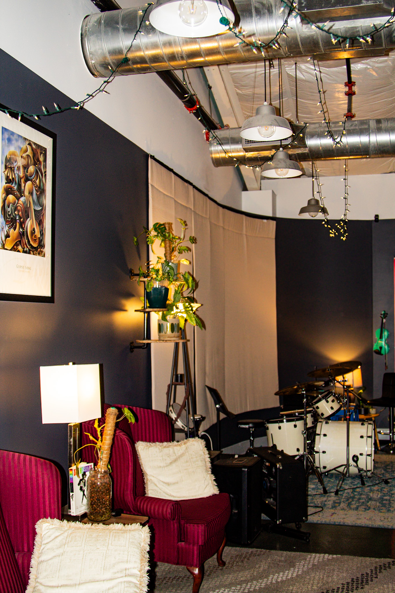The main room set up for a live show - drum kit, red velvet armchairs, and string lights across exposed ductwork