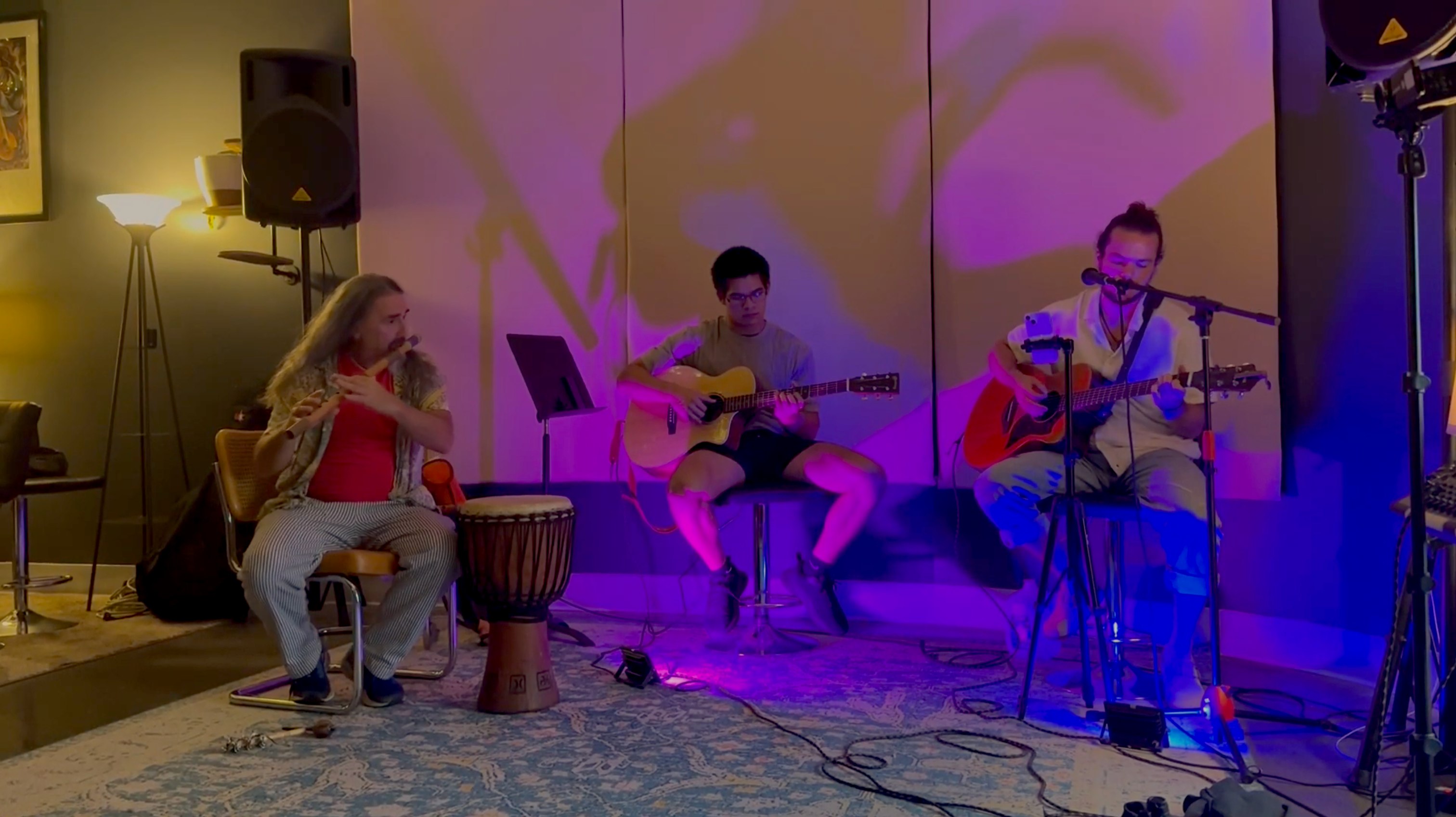 A trio performing live at Soundcheck - a flute and djembe player, an acoustic guitarist, and a singer-guitarist under warm purple stage light