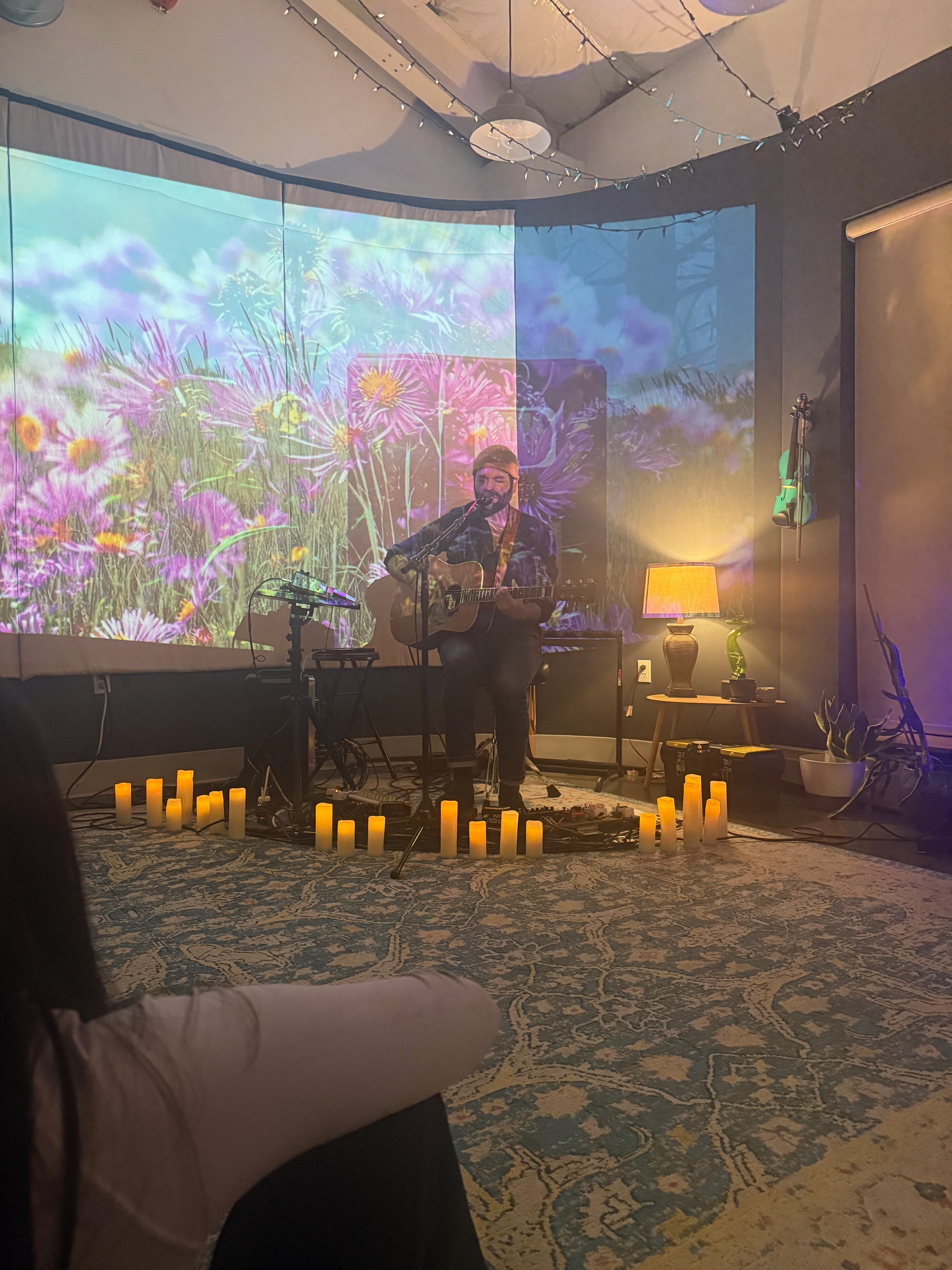 A solo singer-songwriter playing acoustic guitar inside a ring of candles, with a glowing wildflower projection behind them at a Nightcap Session