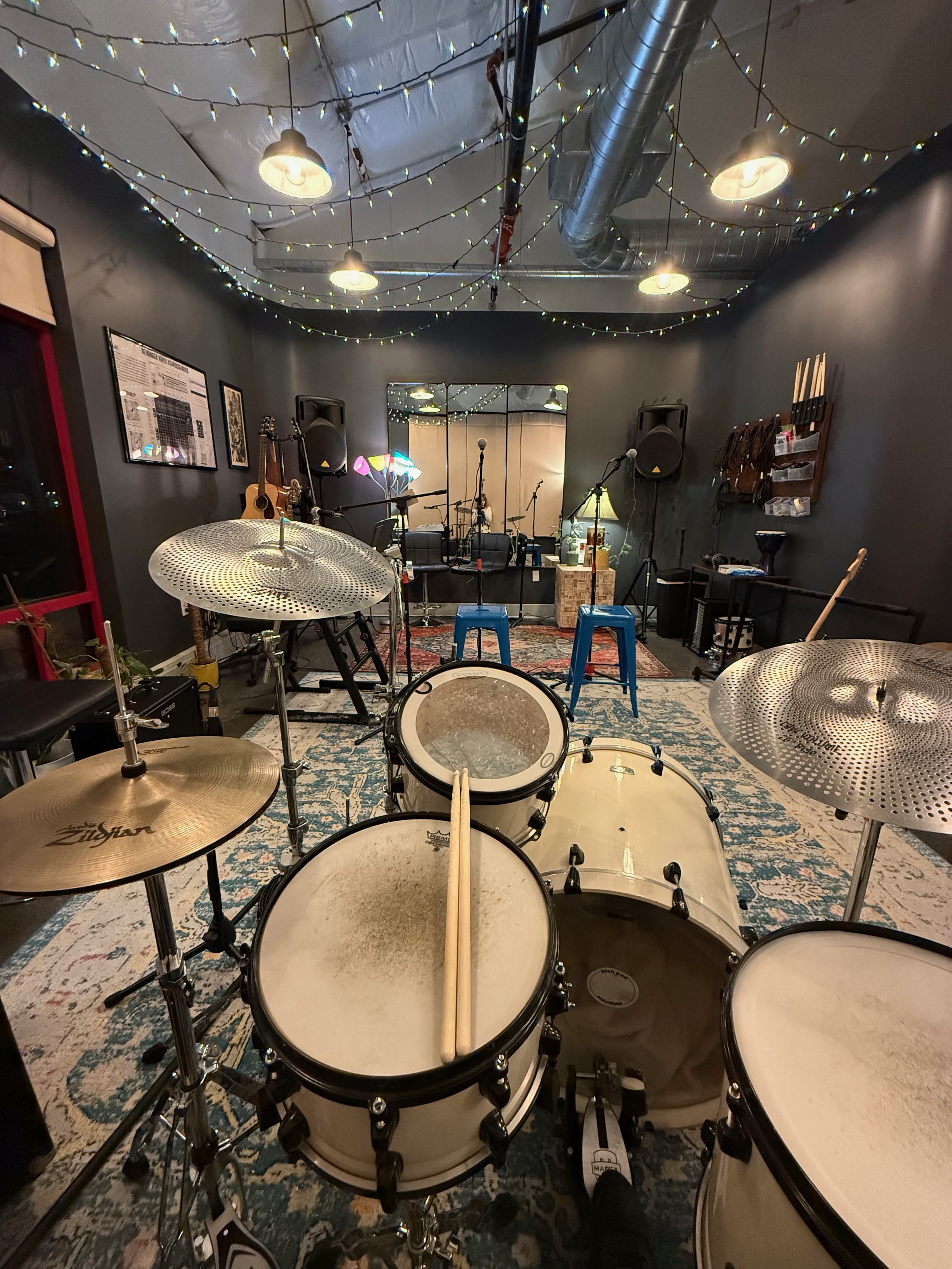 The practice room seen from behind the drum kit - low-volume cymbals, guitars on the wall, PA speakers, and string lights across the ceiling
