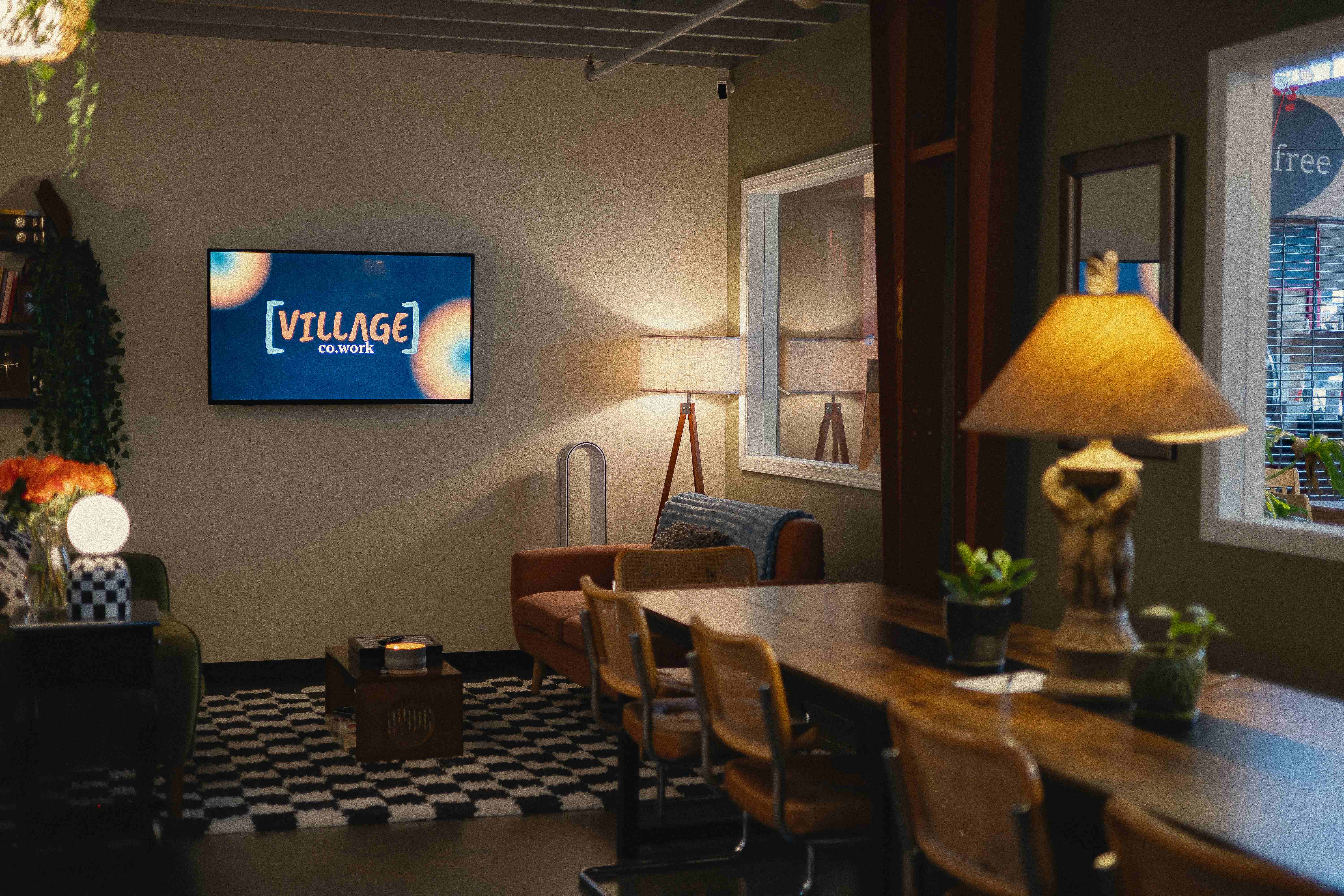 The warm, lamp-lit Villageco.work lounge with a long communal table, sofa, and plants